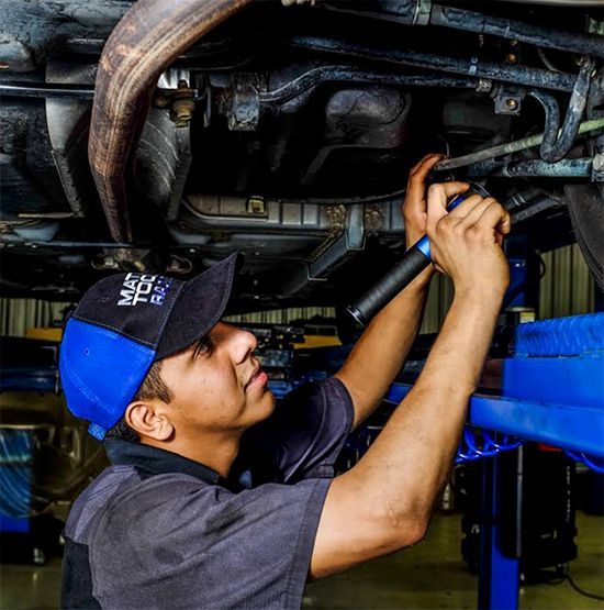 A technician in a blue and black cap uses a flashlight to inspect the undercarriage of a car raised on a lift.