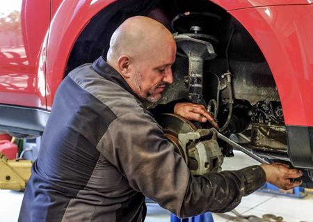 Mechanic working on a car's brakes. Bald man in a gray jumpsuit leaning under the red car, using a wrench.