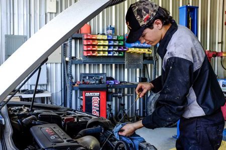 Mechanic checking a car engine in a garage. Wearing a hat and work clothes, with tools and parts visible.