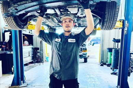 Mechanic under car on lift, in a garage. Wearing work clothes, holding up the car, and smiling.