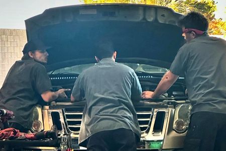 Three mechanics look at a car engine with the hood open. They're wearing work shirts in an outdoor setting.