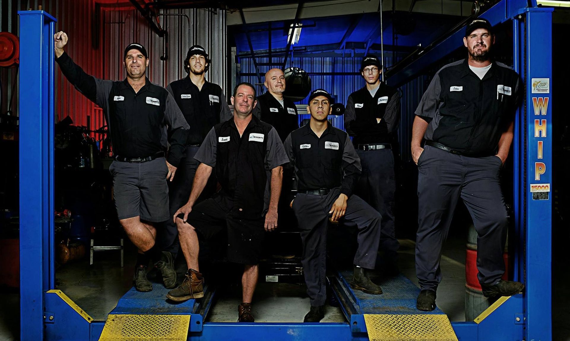 Mechanics in uniforms pose on a car lift in a shop, some leaning.
