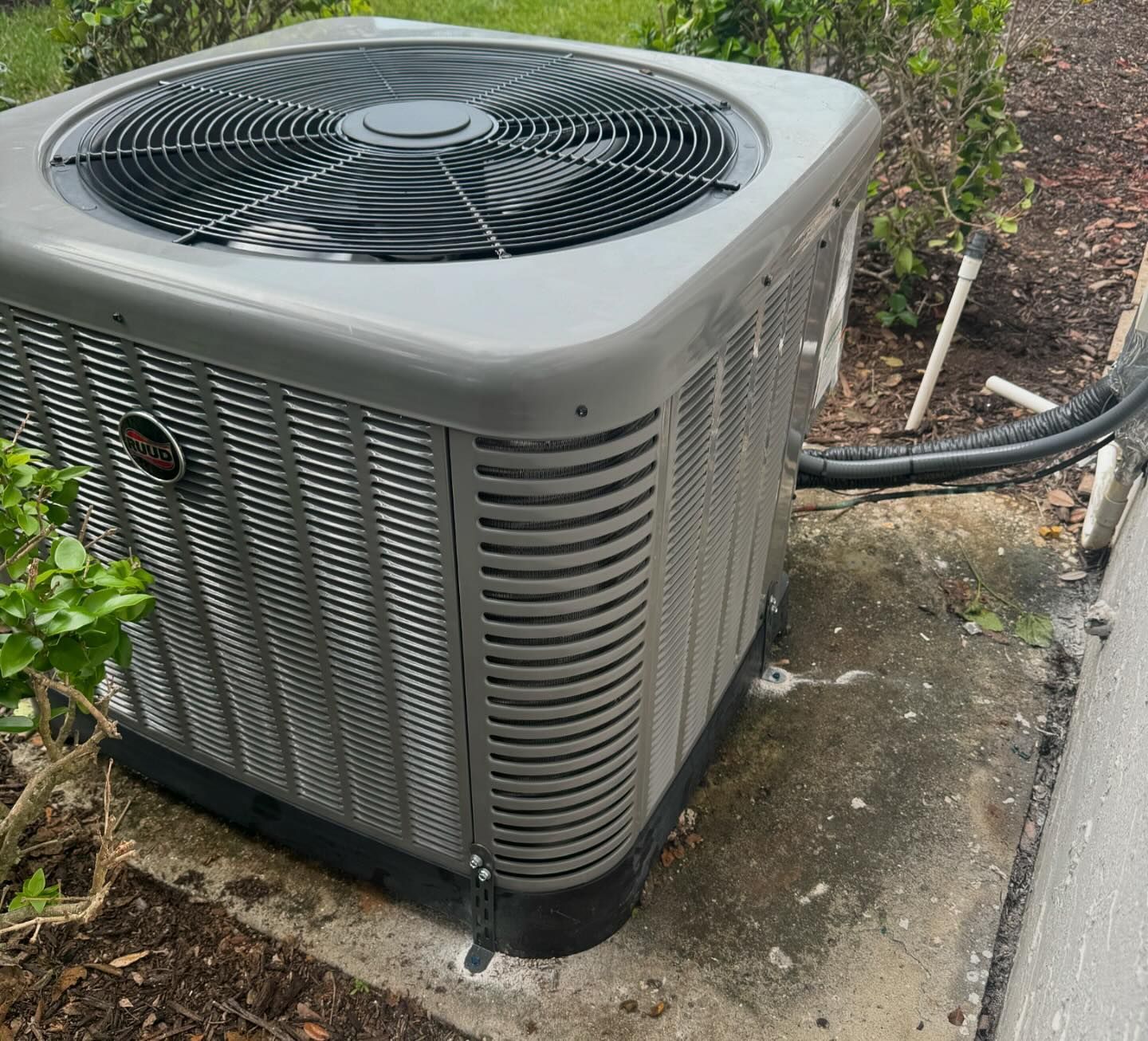 A gray air conditioner is sitting on a sidewalk next to a bush.