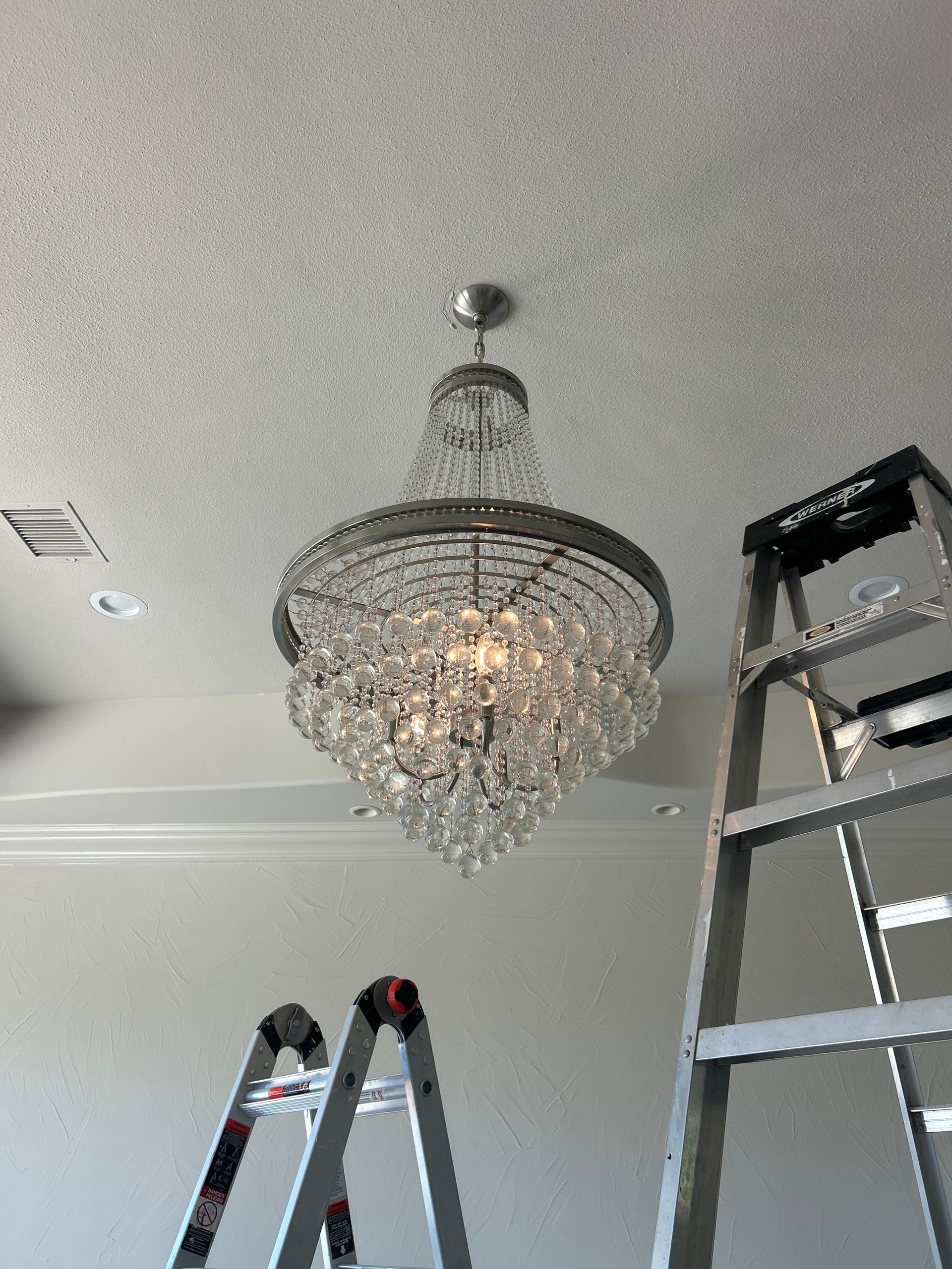Crystal chandelier hanging from a white ceiling with a ladder beside it.