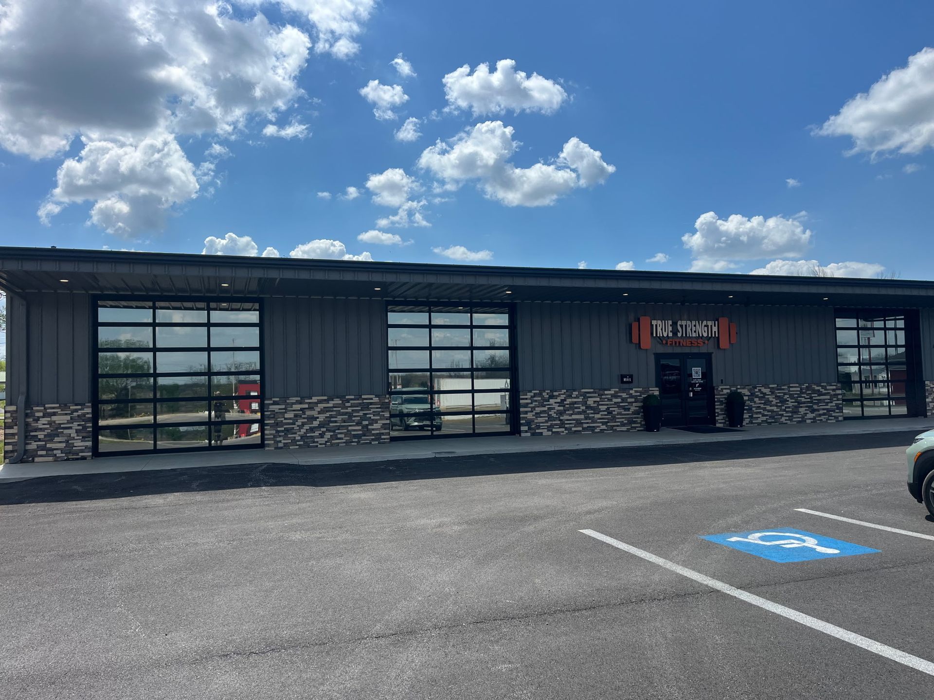 A modern, grey commercial building with large glass garage-style doors, stone accents, and a handicap parking spot.