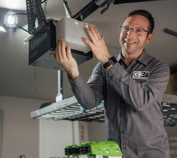 A person in a gray uniform repairing a ceiling-mounted LiftMaster garage door opener in a garage.