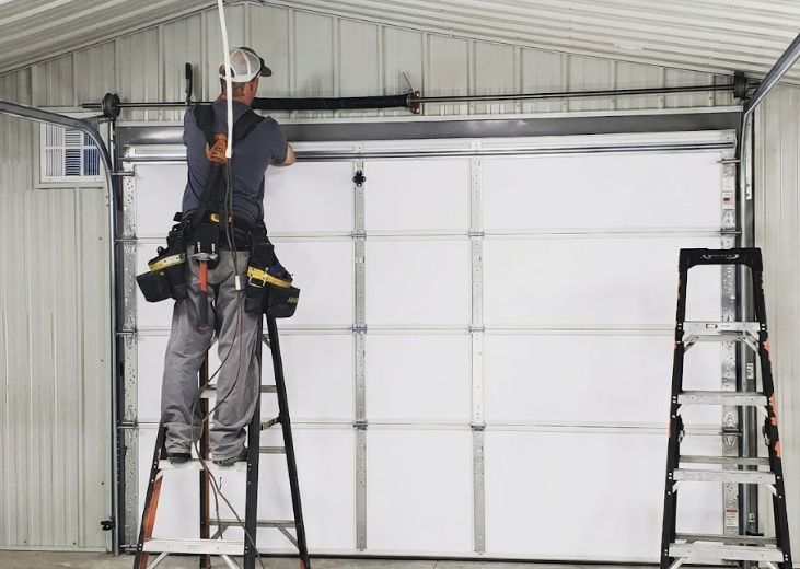 A technician in a hard hat stands on a ladder working on the springs and cables of a white garage door.