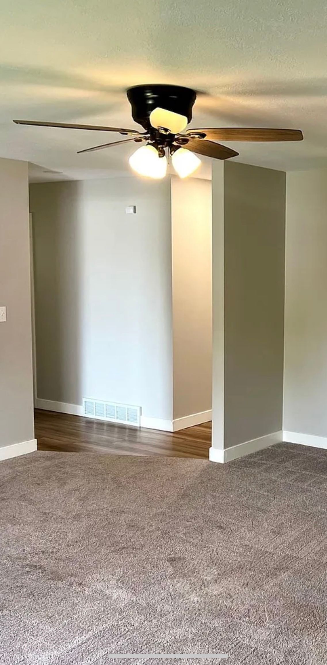 A room with a ceiling fan and a carpeted floor, leading to another area with wooden flooring.