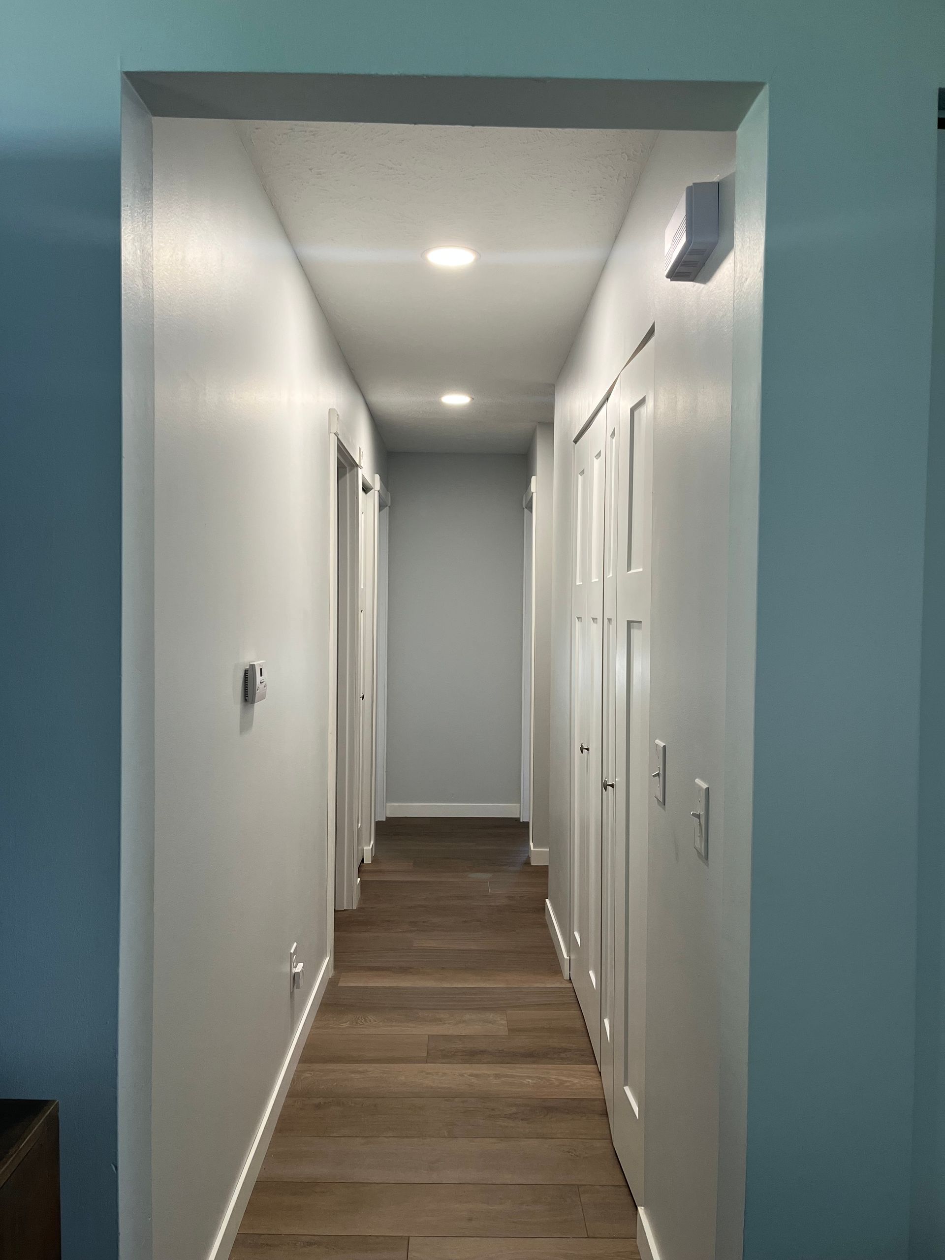Narrow hallway with light gray walls, brown floor, recessed lighting, and white doors.