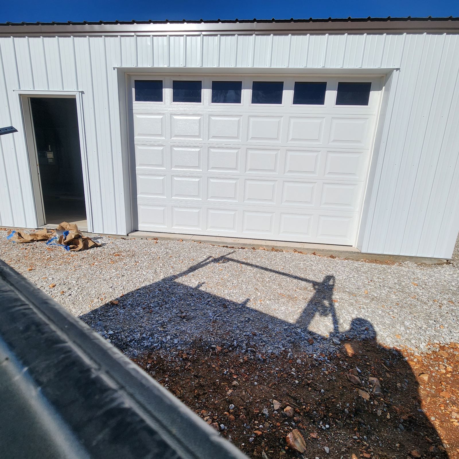 A white metal building with a single-car garage door and a separate side entry door, viewed from a gravel driveway.