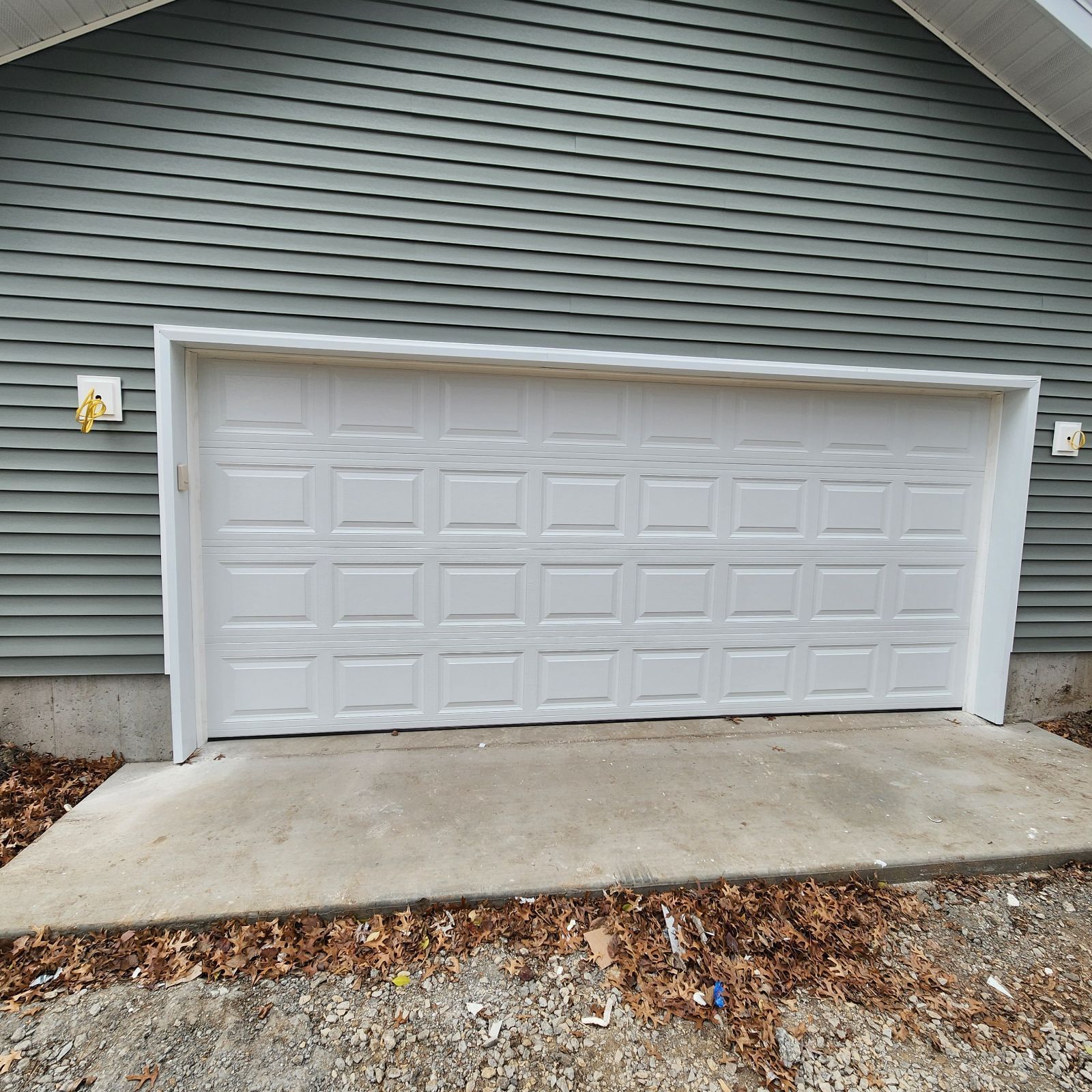 A white garage door set into the green vinyl-sided wall of a house, above a concrete apron and gravel driveway.