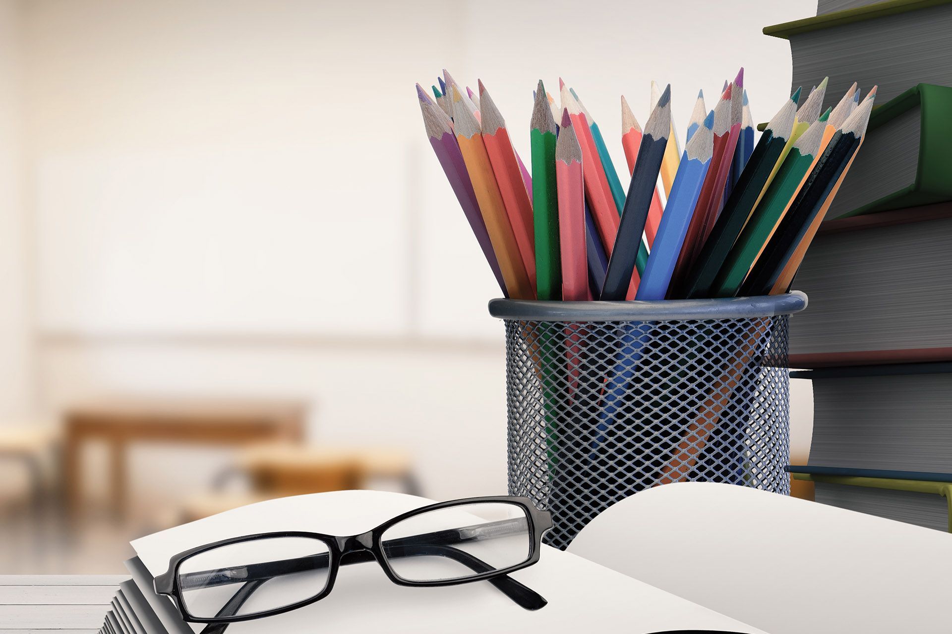 Pencils in a mesh cup, books, and glasses on a desk in a classroom.
