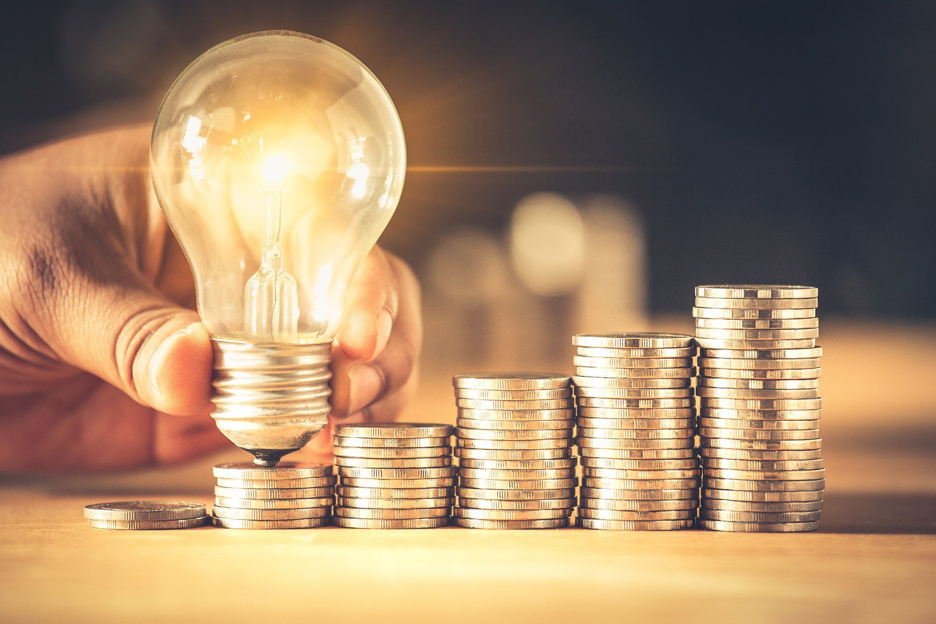 A person is holding a light bulb over stacks of coins.
