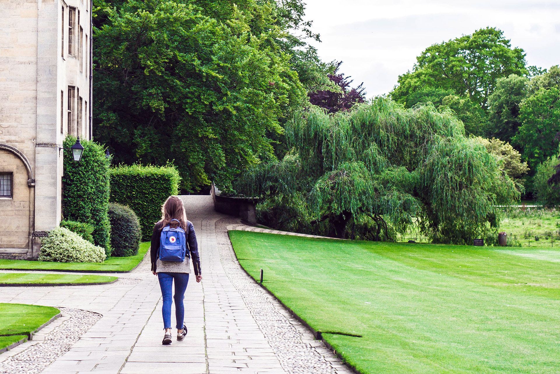 A woman with a backpack walks along a stone path beside a grassy lawn and trees, next to a building.