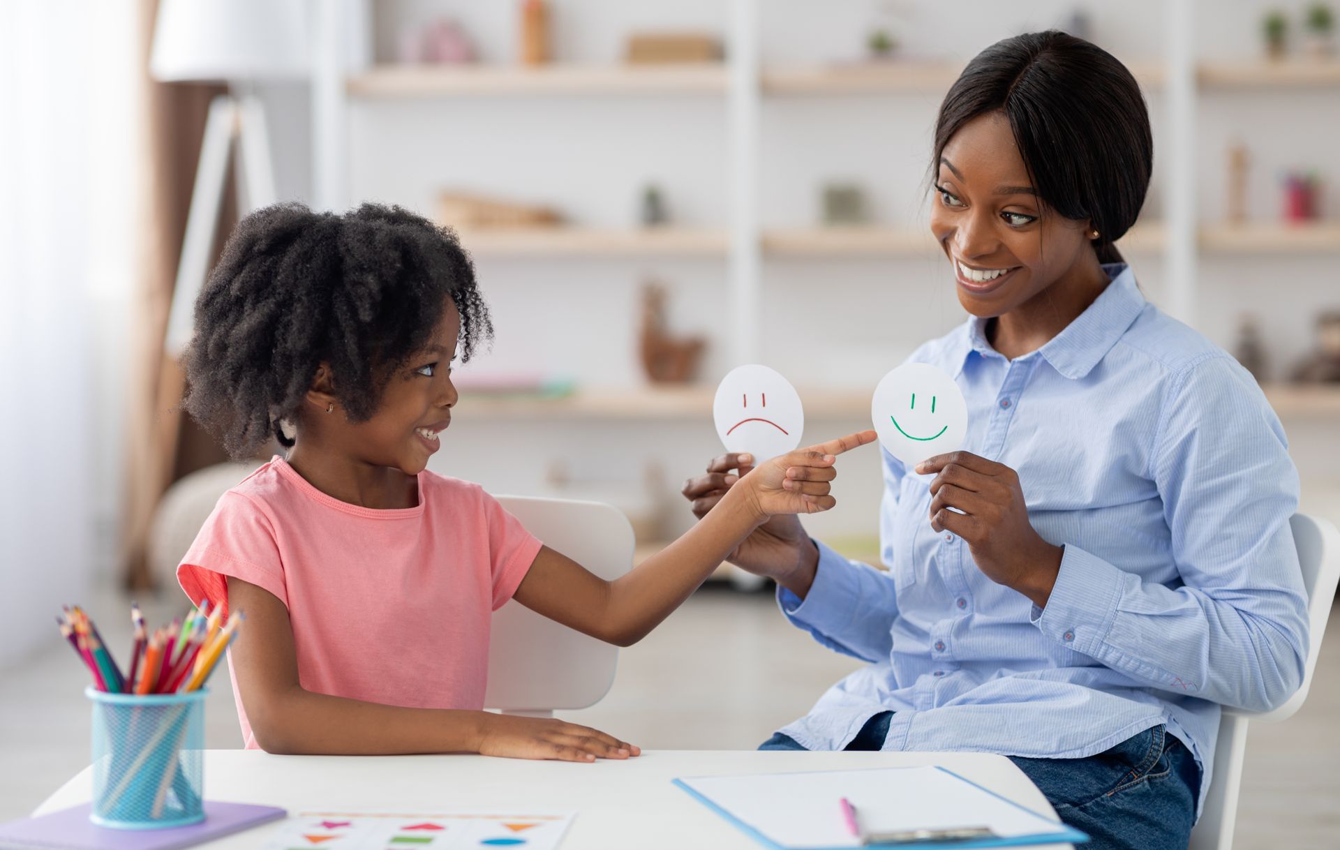 Therapist showing a smiling face card to a child, pointing to the happy face, in a light-filled office.