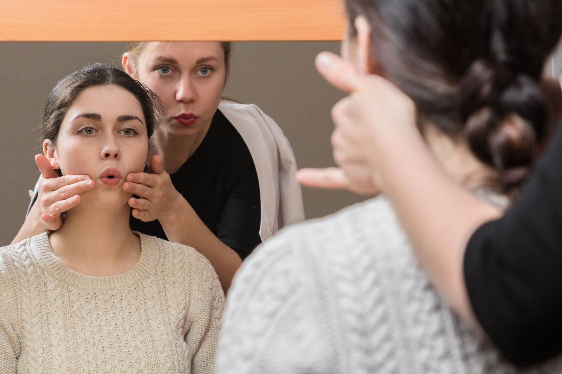 Woman in mirror practicing facial exercises with instructor. Hands gently guide her face.