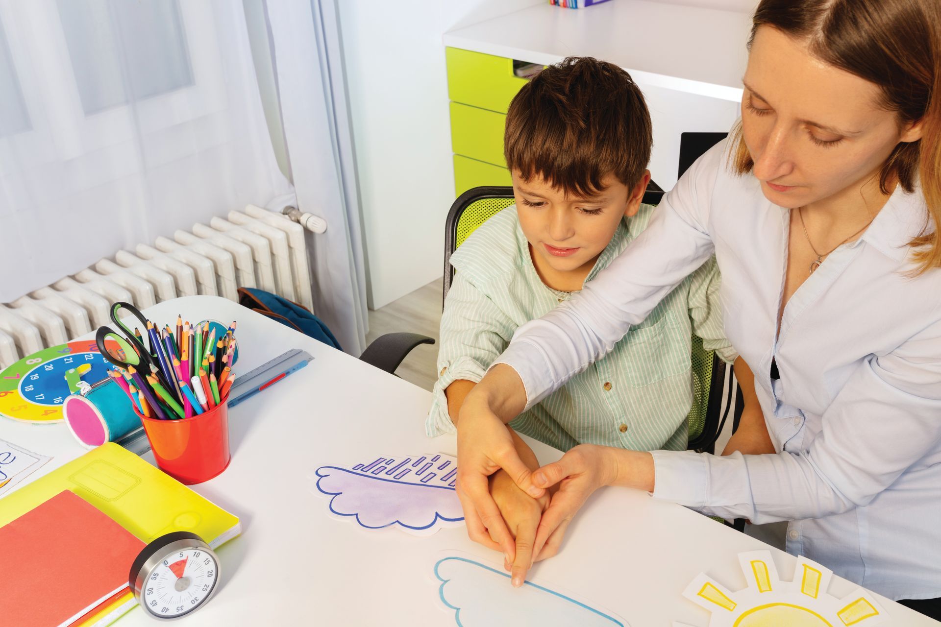 Woman and boy at a table, making a heart shape over a drawing. Tools and art supplies are nearby.