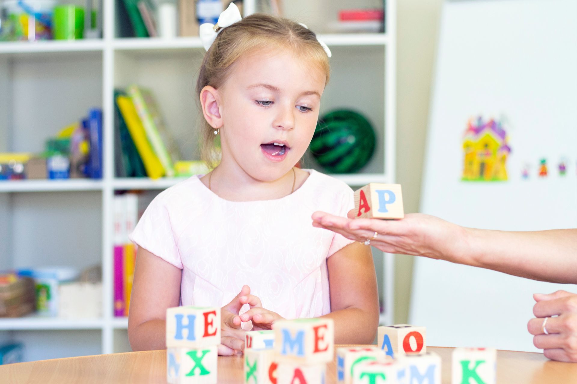 Girl looking at alphabet blocks held by a person. Room with shelves, books, and colorful decor.