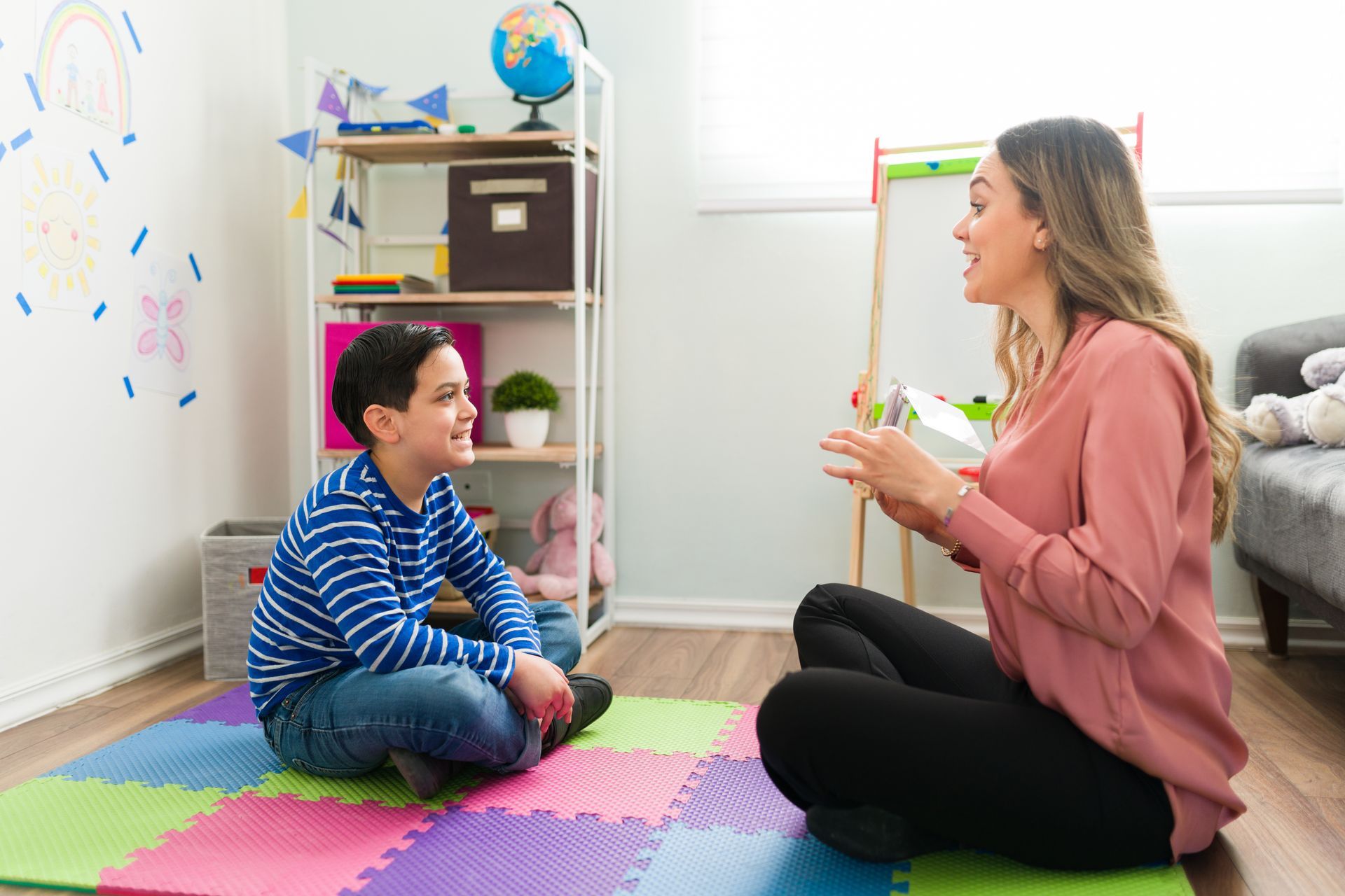 Woman and boy sit on floor, talking in colorful room. Shelves with globe and toys are visible.