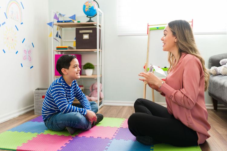 Woman and boy sit on floor, talking in colorful room. Shelves with globe and toys are visible.