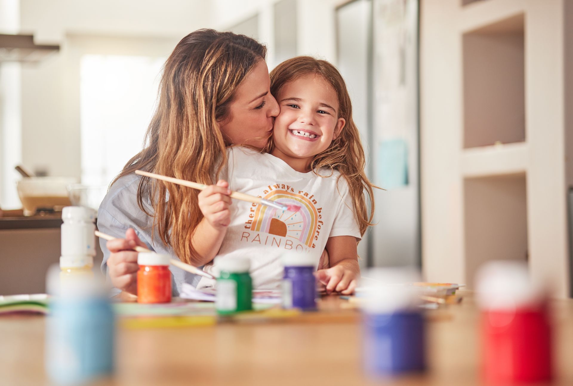 Woman kissing a smiling child holding a paintbrush, surrounded by paint bottles.