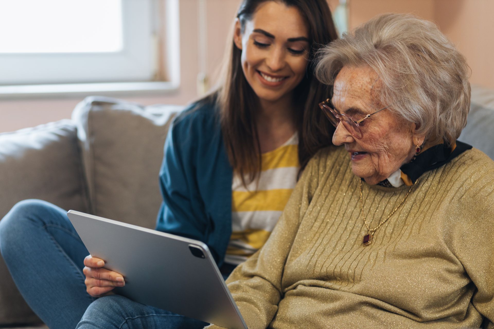 Woman and older person looking at a tablet together, smiling on a couch indoors.