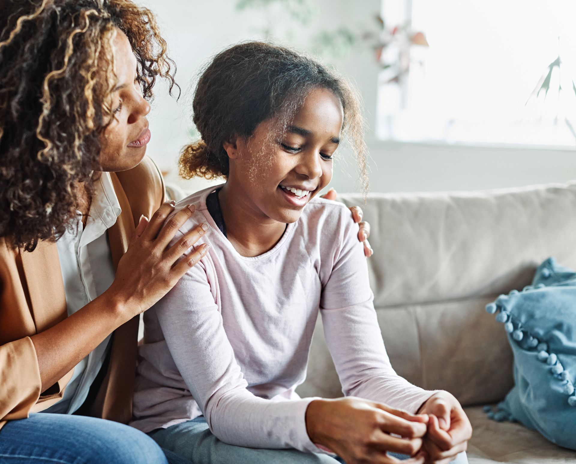 Woman comforts a girl, both sitting on a sofa. Woman has arm around girl's shoulders.