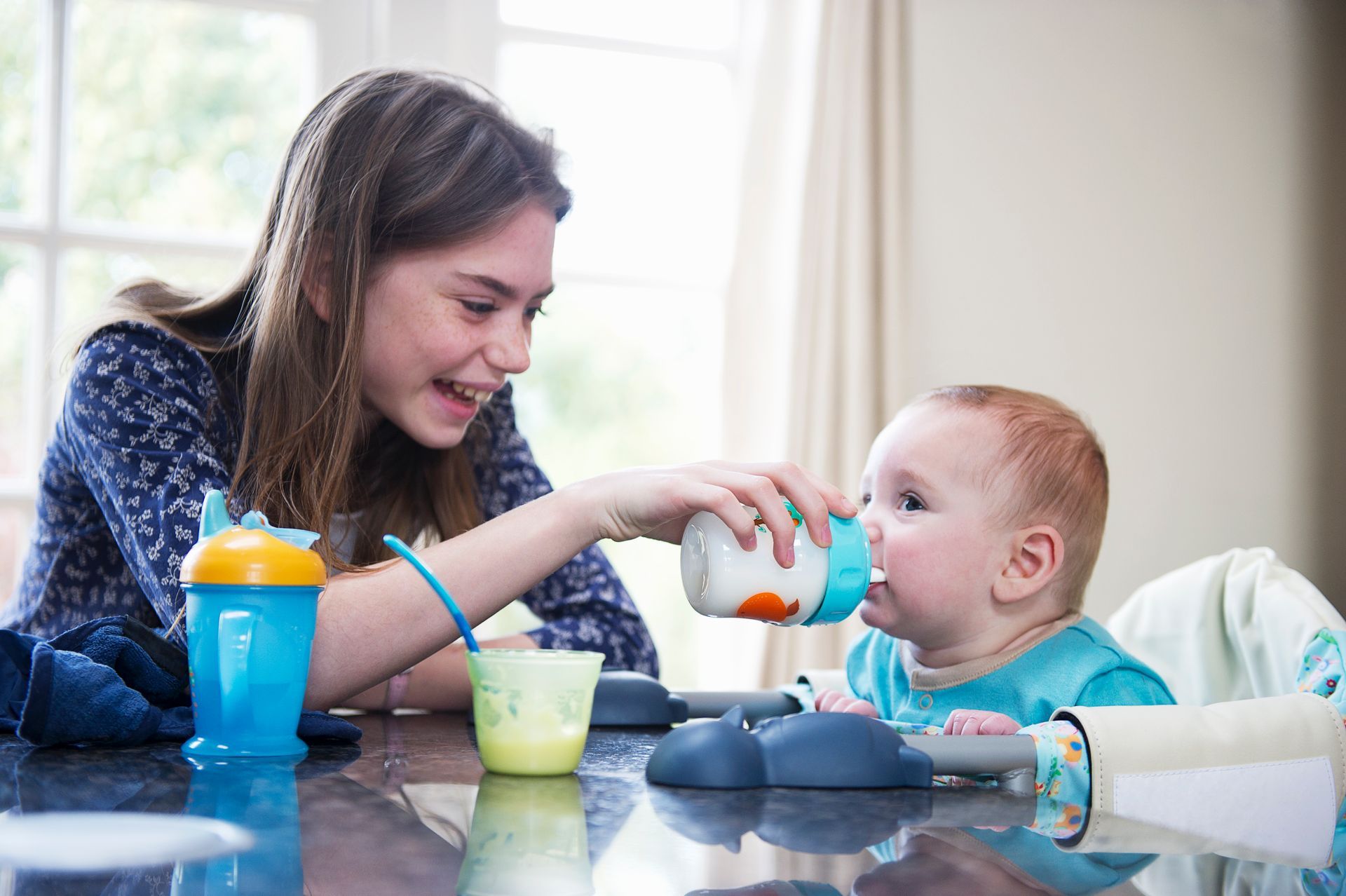 Teen girl smiles, holding a bottle for a baby seated in a high chair at a table.