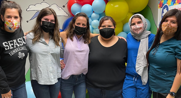 Six people with masks pose for a photo in front of balloons, smiling.