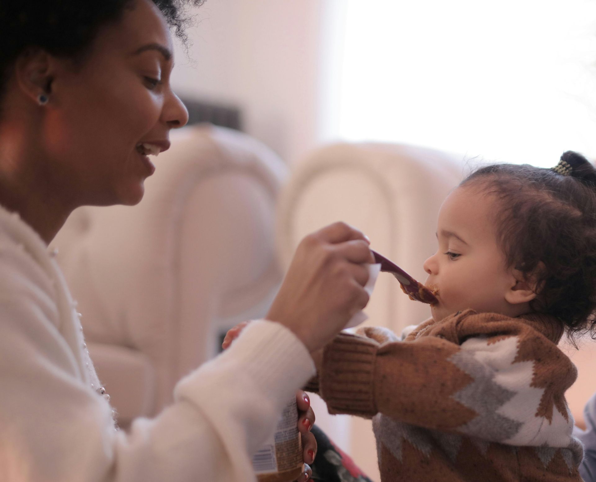 Woman feeding a young child with a spoon. Both are indoors, with soft lighting.