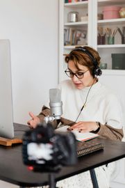 Woman with headphones and glasses speaking into a microphone at a desk, laptop and camera in front.