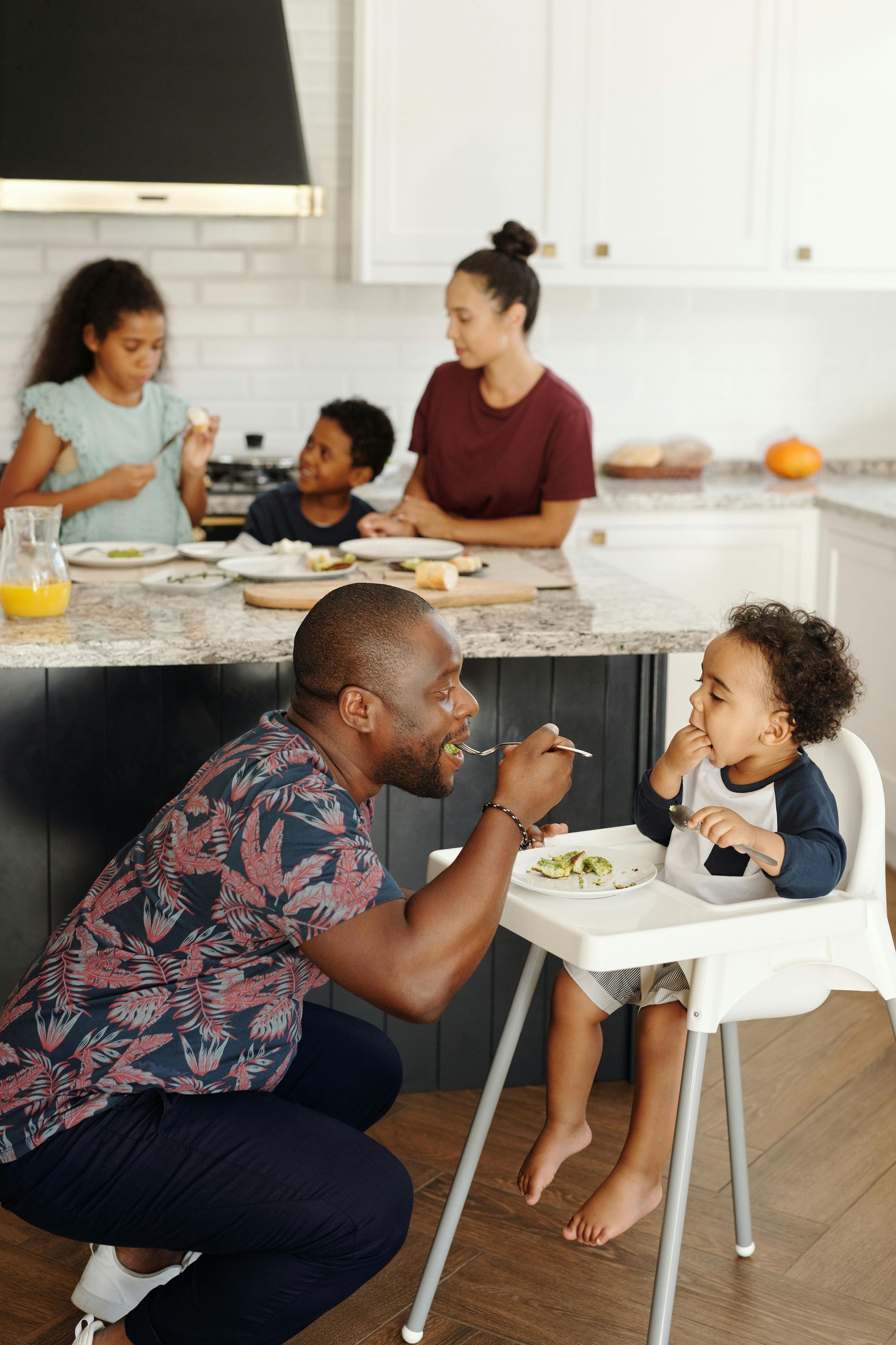 Father feeds toddler in a highchair, family eating at kitchen island.