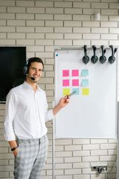 Man with headset points at a whiteboard with sticky notes in front of a brick wall.