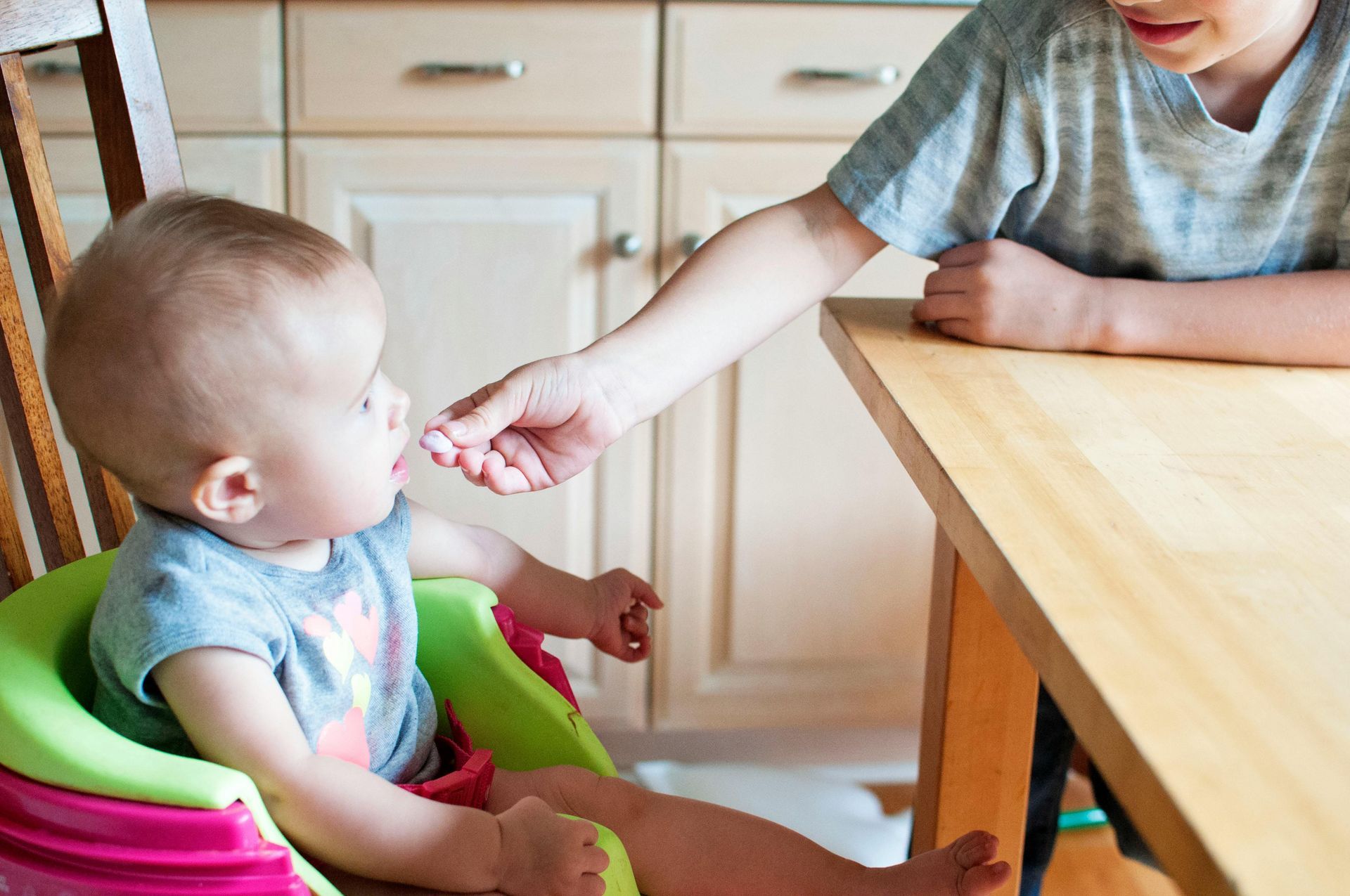 A child feeding a baby in a high chair at a wooden table in front of kitchen cabinets.