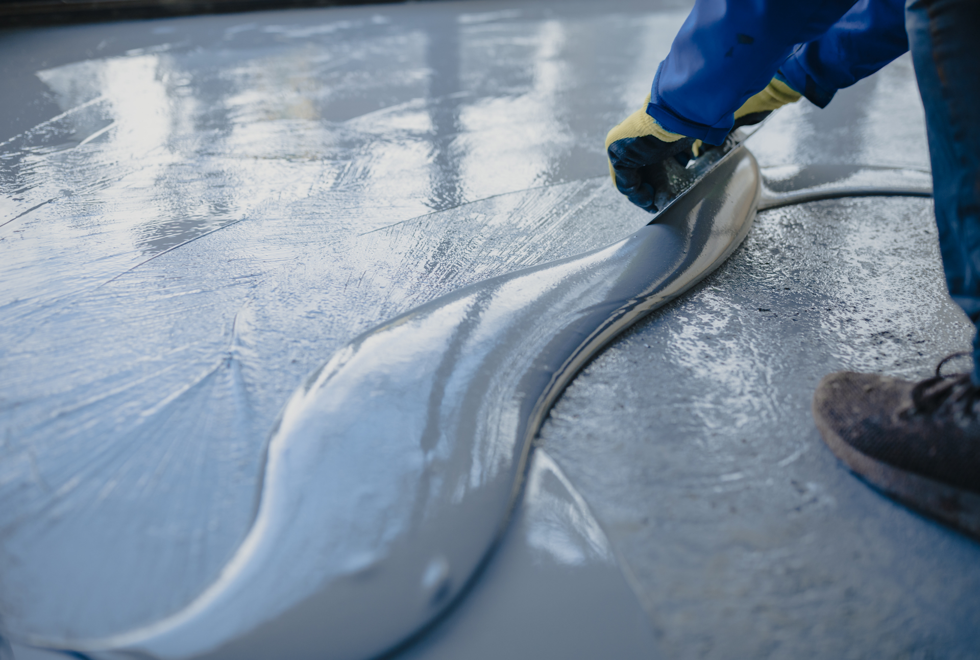 Person using a trowel to spread wet concrete on a smooth, gray surface.