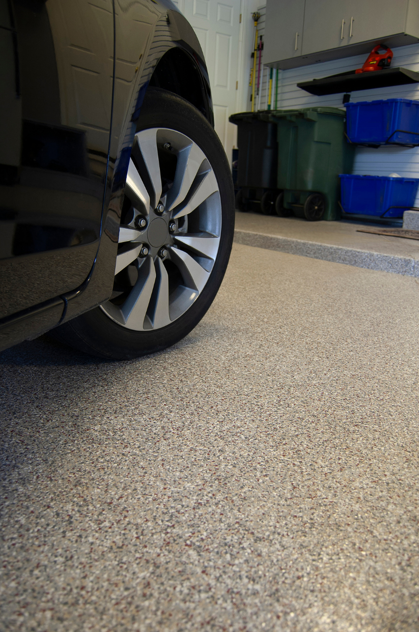 Black car wheel on a speckled garage floor. Trash cans and storage containers in the background.