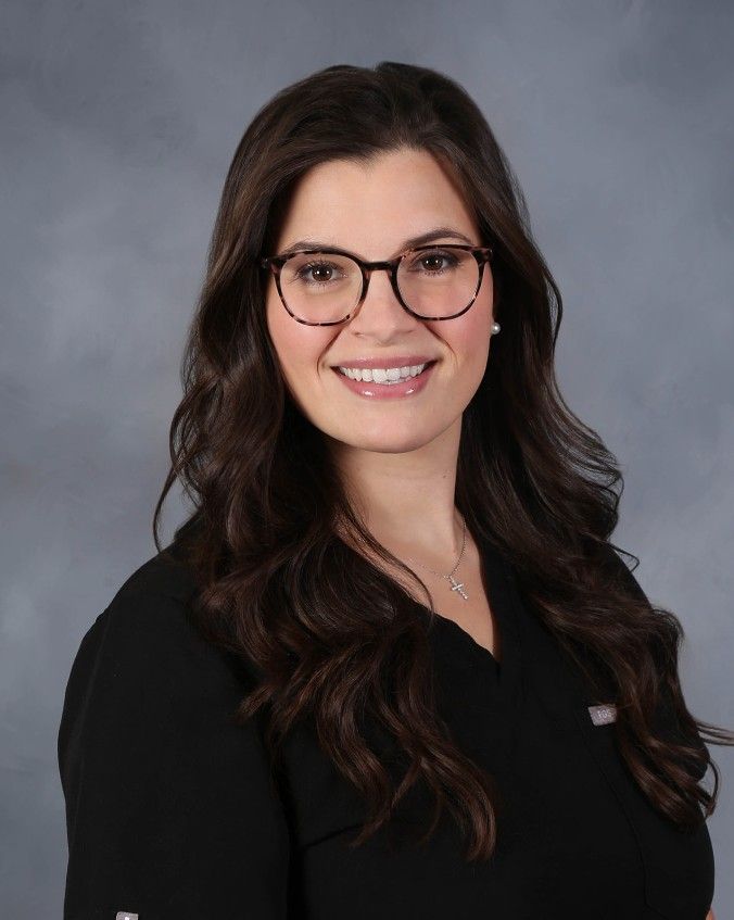 Woman with long brown hair smiles, wearing a black shirt against a gray background.