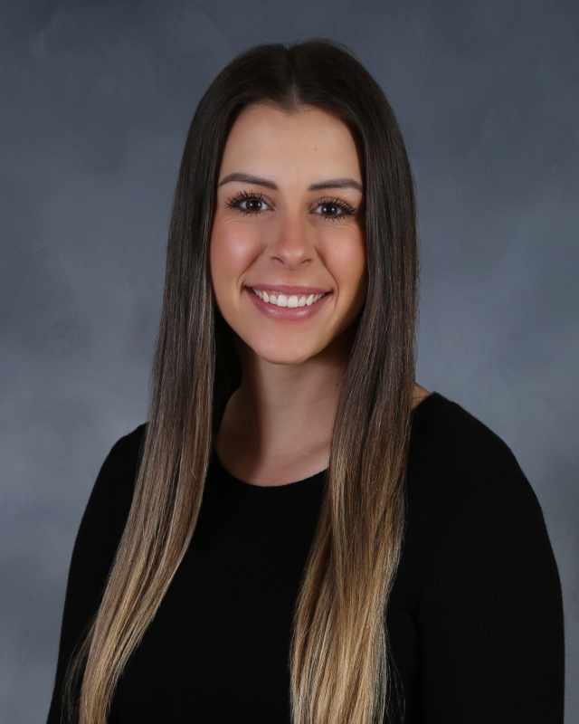 Woman with long brown hair smiling at the camera. Wearing a black shirt, against a grey background.