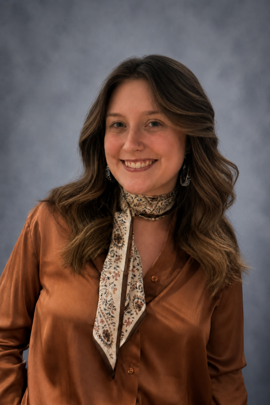 Smiling person in a brown blouse and patterned scarf against a gray studio backdrop. Kenzi