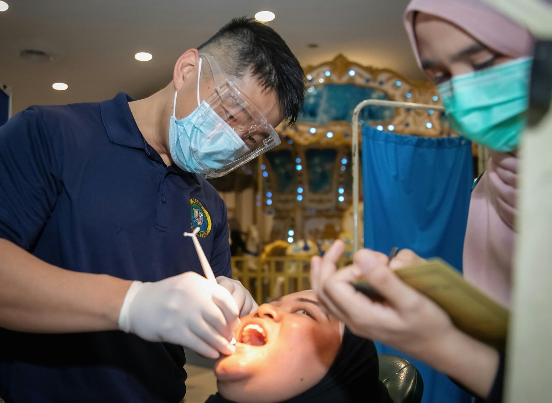 Dentist examining a patient's mouth. Assistant observes. Indoors, dental setting.