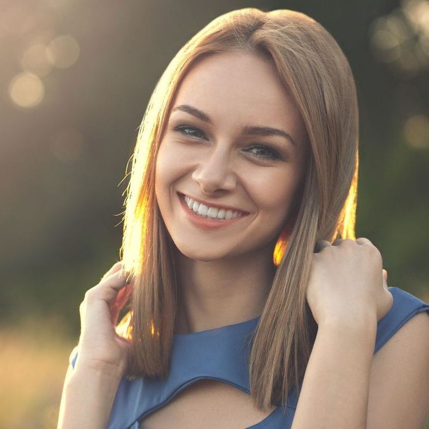 Woman with blonde hair smiles, hands near her face, in a sunlit outdoor setting.
