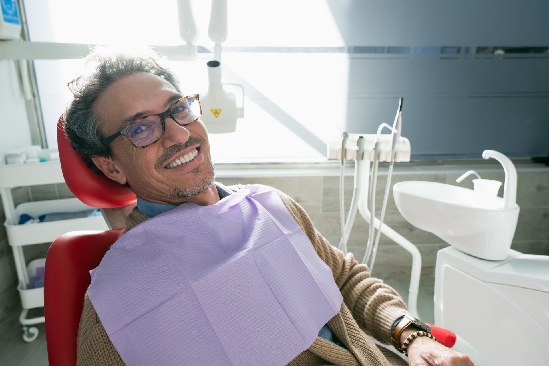 Man in dental chair, smiling at camera; wearing glasses and bib, in a bright, modern dental office.