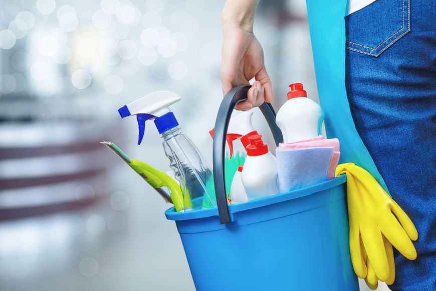 Person carrying blue cleaning bucket with spray bottles, gloves, and detergents.