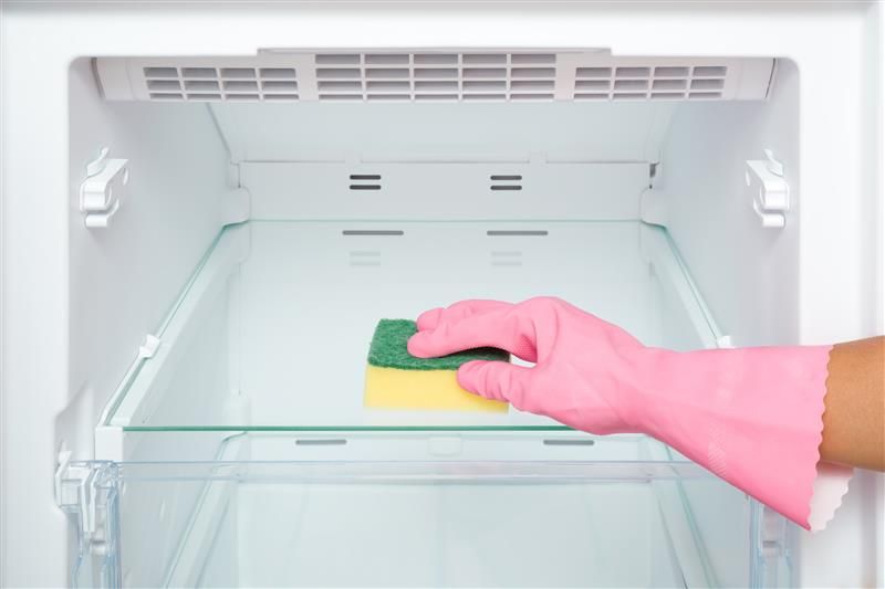 A gloved hand cleans the empty refrigerator shelf with a sponge.