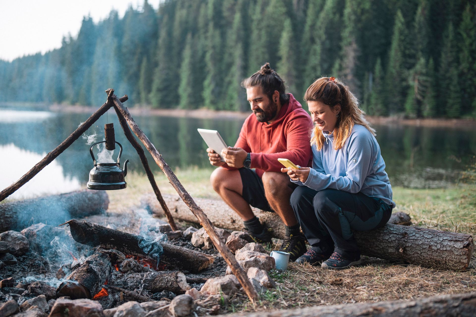 a man and a woman are sitting next to a campfire looking at a tablet .