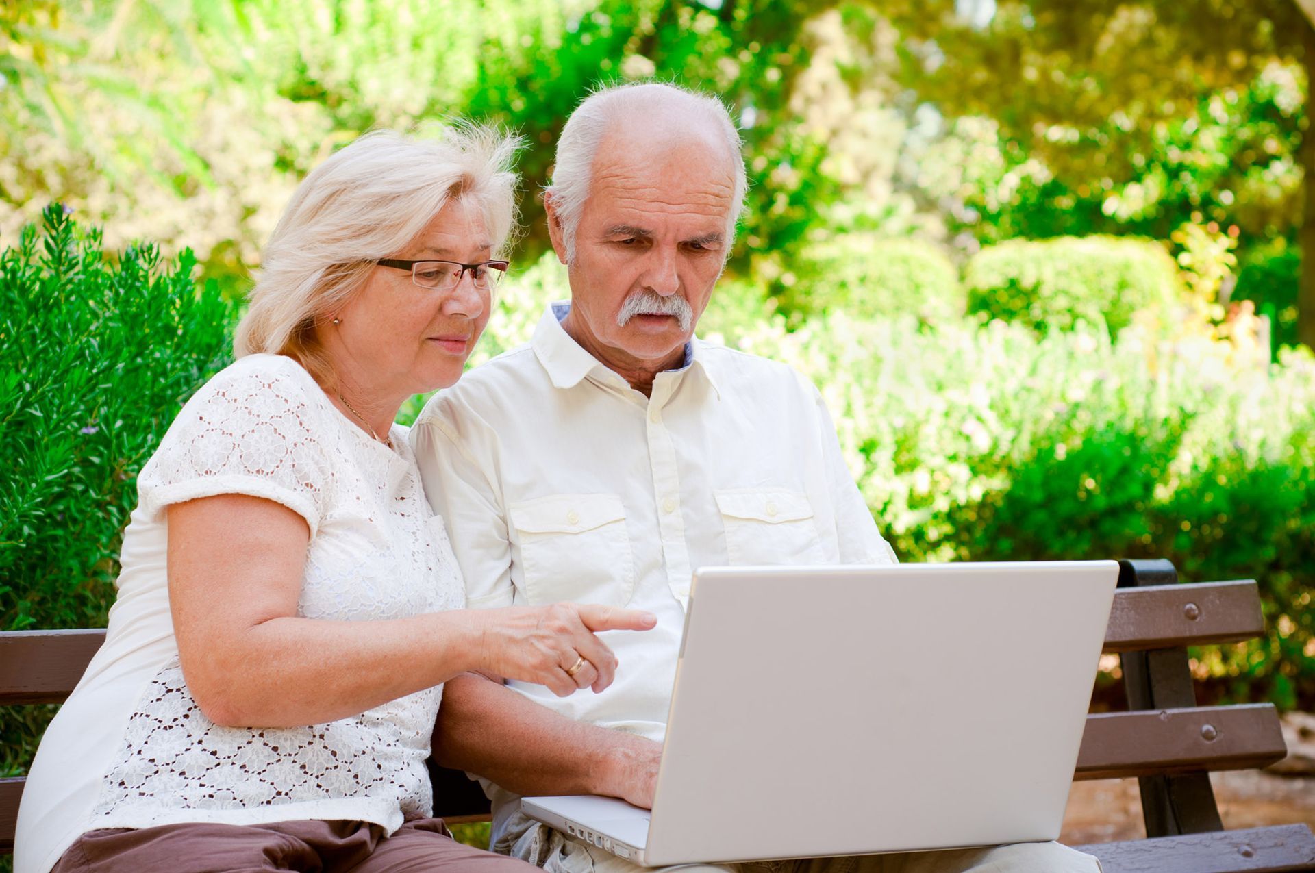 an elderly couple is sitting on a bench looking at a laptop computer