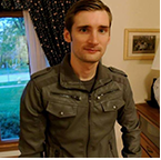 A man in a jacket is standing in front of a window in a living room.