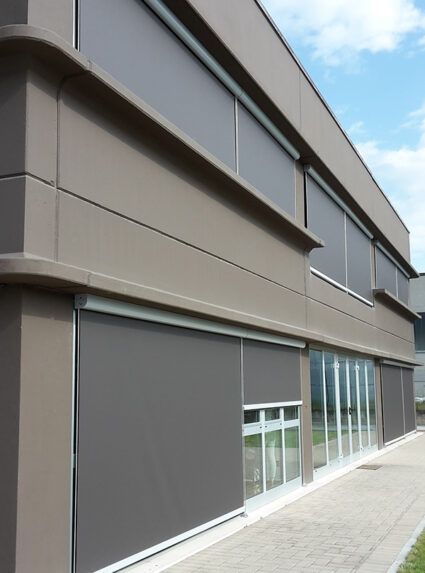 Edificio grigio con tende parasole grigio scuro che coprono le finestre. Cielo azzurro visibile.