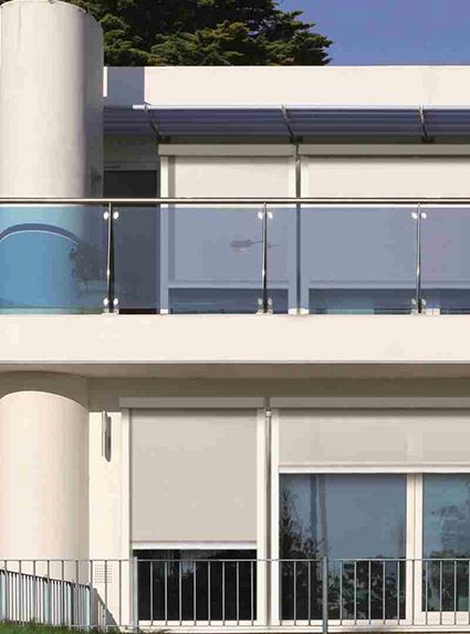 Moderno edificio bianco con balconi in vetro, persiane grigie e cielo azzurro.