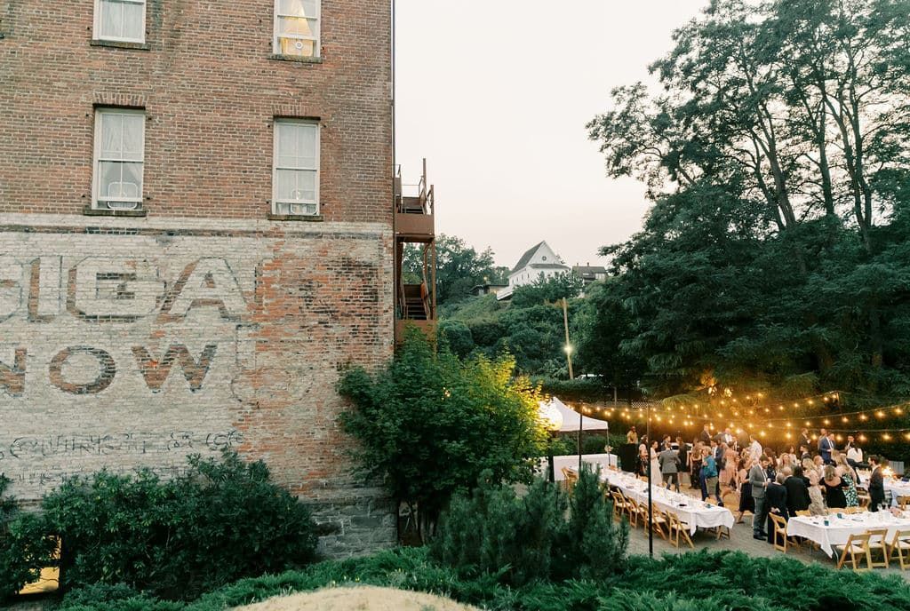 A group of people are sitting at long tables in front of a brick building.